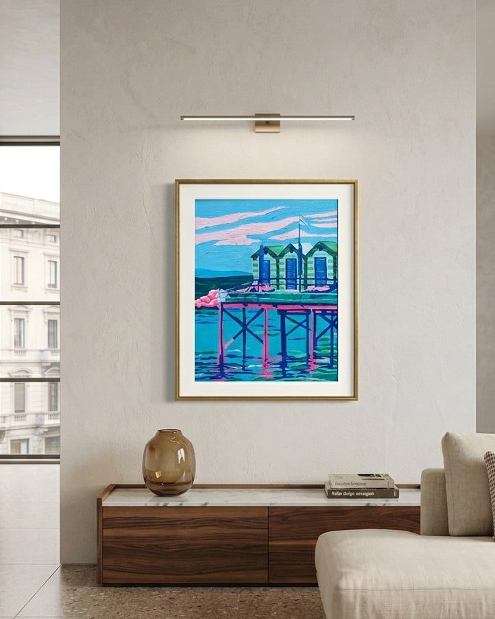 A framed pop art print of a pier scene hangs on a light gray wall in a modern living room setting with a beige sofa and a wooden console table.