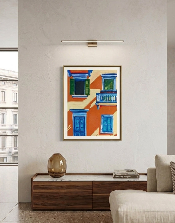 A framed art print depicting colorful Mediterranean-style building facades with blue doors and windows, displayed on a light beige wall in a modern living room setting.