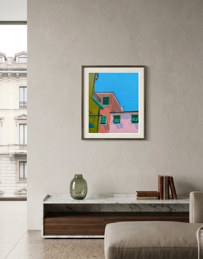 A framed colorful art print depicting pink and green buildings under a bright blue sky, displayed on a textured beige wall in a modern living room setting.