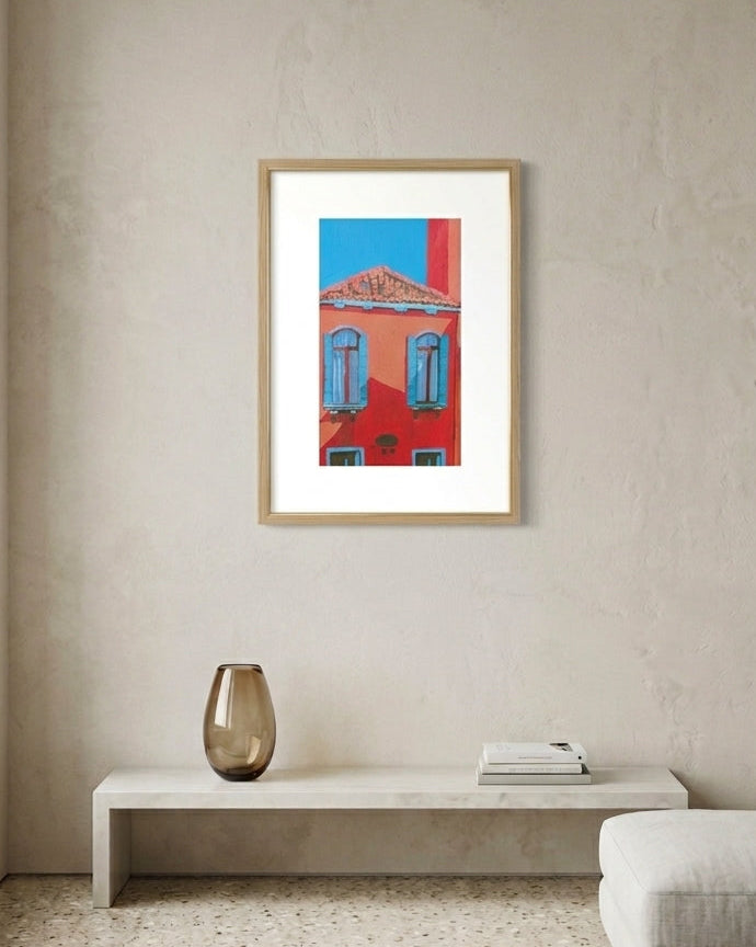 A framed art print depicting a red building facade with blue shutters under a bright blue sky, displayed on a textured beige wall in a modern living room setting above a low white bench.